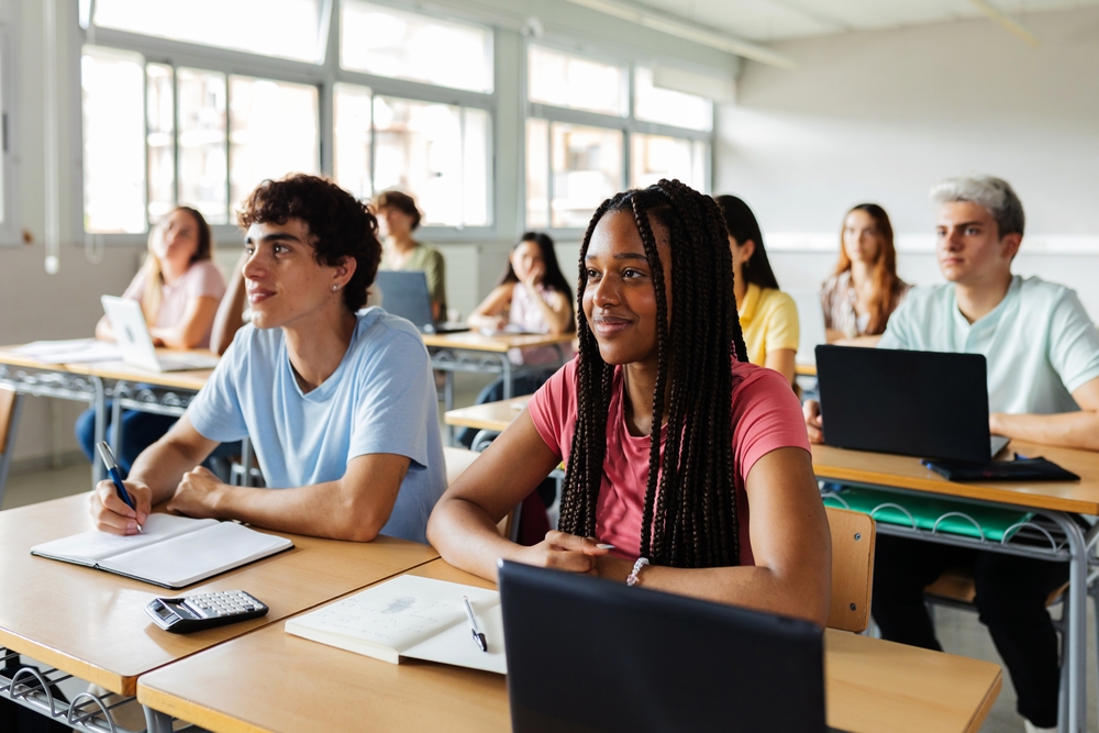 A group of diverse students attentively listening in a classroom. They are seated at desks with notebooks and laptops, facing the front of the room. Sunlight streams through large windows, creating a bright and engaging atmosphere.