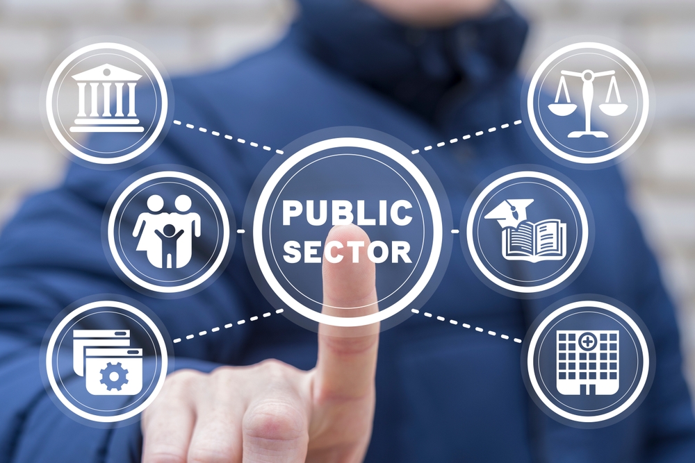 A person pointing at a digital interface displaying the term "Public Sector" at the center, surrounded by interconnected icons. The icons represent various aspects of public services, including a government building, family and social services, legal balance scales, education, healthcare, and administrative processes. The image emphasizes the interconnected nature of public sector functions and services.